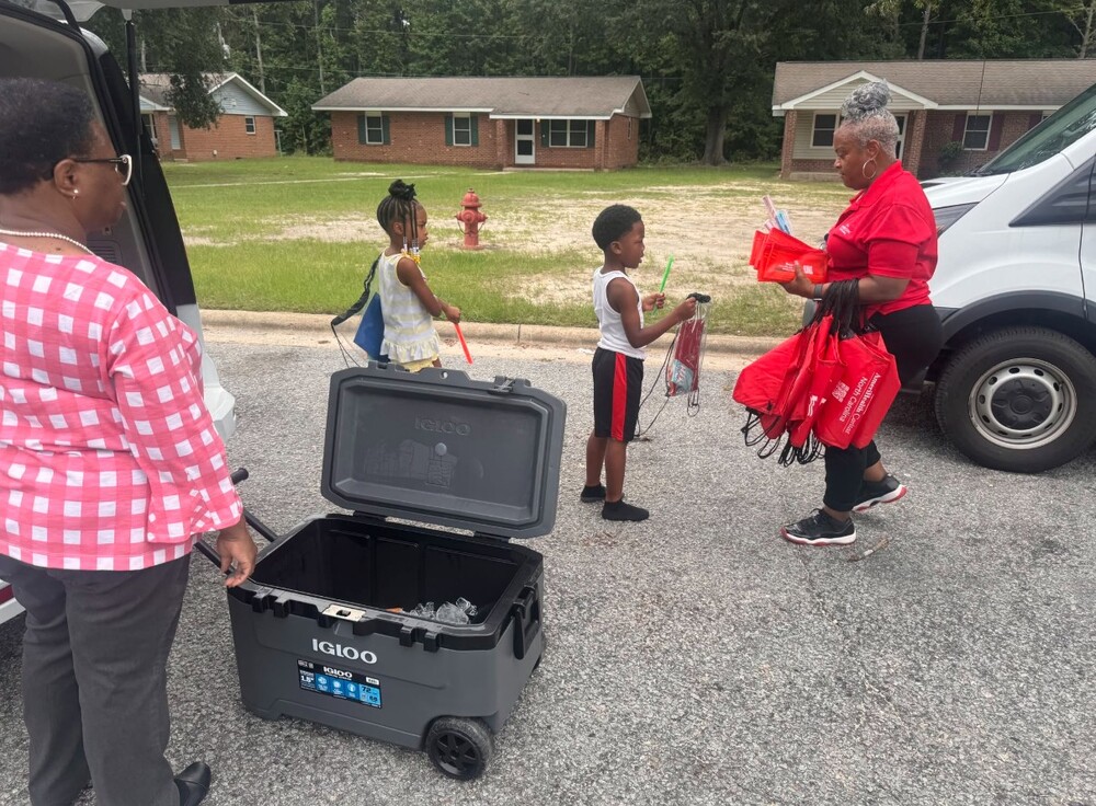 A woman standing by a cooler watching another woman handing out school supplies to a young boy.
